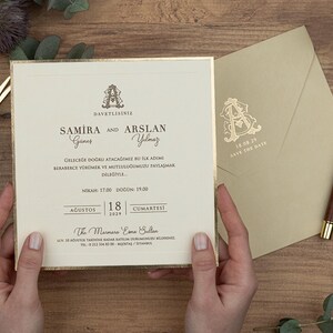 May include: An ivory wedding invitation with gold edging, held by hands. The invitation features elegant script and the names "Samira and Arslan." An envelope and pen are also visible. The date "August 18, 2019" is printed on the invitation.