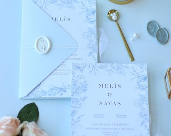 May include: A light blue wedding invitation with a floral design and the text "MELIS & SAVAS" in a dark blue font. The invitation includes the date, time, and location of the wedding. The invitation is decorated with a white wax seal.