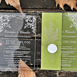 Puede incluir: Una invitación de boda de acrílico transparente con un sobre verde. La invitación presenta un diseño floral blanco y el texto "Carmine e Claudia" y "Annunciano con felicita il loro matrimonio". El sobre presenta un diseño floral verde y el texto "e Claudia" y "cita il loro matrimonio".