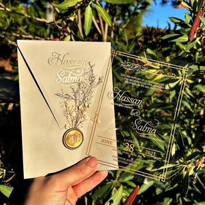 Puede incluir: Invitación de boda con los nombres "Hassan" y "Salma" en letras doradas. La invitación incluye un sobre color crema con un sello de cera y una tarjeta acrílica transparente con los detalles del evento. La fecha es el 28 de junio de 2025.