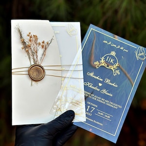以下が含まれることがあります： 白いカードにドライフラワー、金のシール、透明なオーバーレイが付いた、エレガントなウェディング招待状セット。 金色のカリグラフィーとイベントの詳細が記載されたネイビーブルーの招待状カードも含まれています。