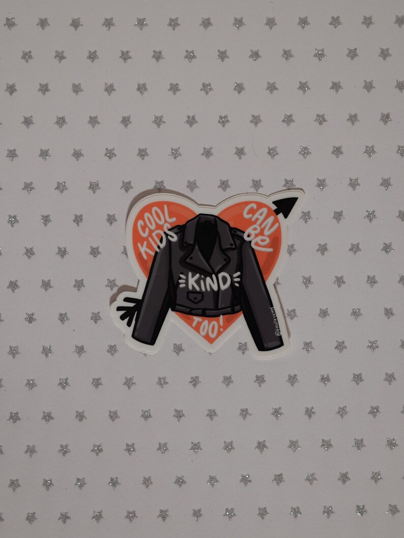 May include: A red heart-shaped sticker with a black leather jacket and the text "Cool Kids Can Be Kind Too!" inside. The sticker is on a white background with silver glitter stars.