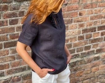 Puede incluir: Una blusa de lino gris oscuro con cuello de volantes y mangas cortas. La blusa se combina con pantalones de lino blancos.
