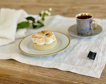 May include: A white linen placemat with a gold rimmed plate and a lemon meringue tart on top. A cup of coffee and a saucer are on the right side of the placemat. The placemat has a small black tag with the word "Linen" on it.