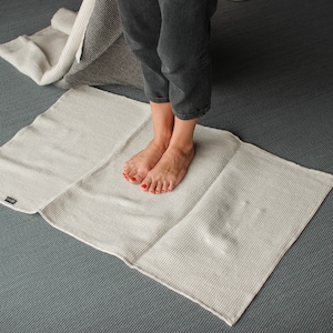 Op de afbeelding: Een lichtbeige geweven badmat is gedeeltelijk uitgevouwen op een grijze vloer. De mat is rechthoekig met een gestructureerd oppervlak. Een tweede opgevouwen mat en de benen van een persoon zijn zichtbaar op de achtergrond.