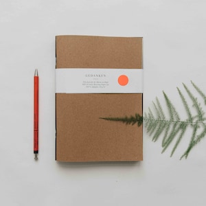Puede incluir: Un cuaderno marrón con una etiqueta blanca que dice "GEDANKEN" y un pequeño círculo naranja. Un bolígrafo rojo y un helecho verde están sobre una superficie blanca.