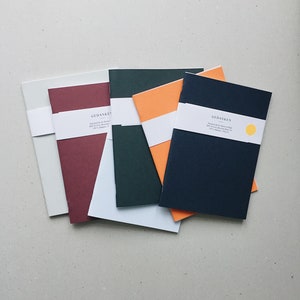 Könnte beinhalten: Ein Stapel von fünf Notizbüchern mit verschiedenen farbigen Umschlägen. Die Umschläge sind weiß, rot, dunkelgrün, orange und dunkelblau. Die Notizbücher haben den Schriftzug "Gedanken" auf dem vorderen Umschlag.