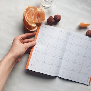 Könnte beinhalten: Ein offener orangefarbener Planer mit einem monatlichen Kalendarium. Der Planer hat einen leuchtend orangenen Einband und die Seiten sind weiß. Der Planer ist für den Monat Januar geöffnet.