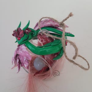 May include: A glass ornament featuring a green dragon with red accents and purple flowers. The dragon is wrapped around the glass, with a twine bow and pink feathers. The ornament has a whimsical, fantasy theme.