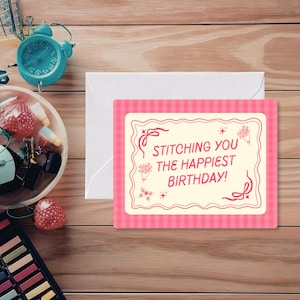 May include: A pink and white birthday card with the text "Stitching You The Happiest Birthday!" sits on a wooden surface next to a white envelope. The card has a pink gingham border and decorative floral elements.