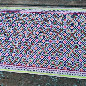 May include: A white fabric placemat with a geometric pattern in shades of red, green, blue, and pink. The pattern is made up of small squares and crosses.