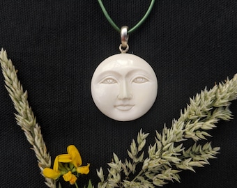 Peut inclure: Un pendentif en os blanc sculpté avec un visage de lune, suspendu à un collier en cordon vert. Le pendentif est sur un fond noir avec des herbes séchées et des fleurs jaunes.