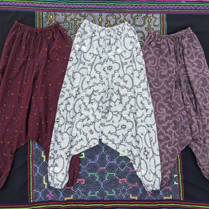 May include: Three pairs of harem pants in varying colors: burgundy with small yellow and blue accents, white with a black geometric pattern, and a mauve color with a swirling design. The pants are displayed on a black patterned textile.