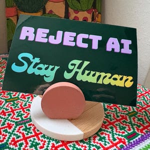 May include: A rectangular dark green sign with the text REJECT AI in light purple and Stay Human in a gradient of light blue, green, and yellow. The sign is held by a circular stand featuring a pink semi-circle on a white and wood base.