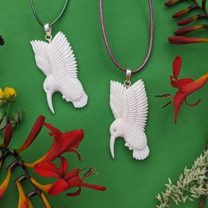 May include: Two white carved hummingbird pendants on brown leather cords. The pendants are hanging against a green background with red flowers.