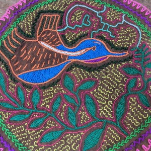 Può includere: Un arazzo da parete circolare ricamato con sfondo marrone. Il disegno presenta un uccello blu e arancione con un lungo becco, circondato da foglie verdi e fiori rosa. Il bordo è un motivo a zig-zag rosa e viola.