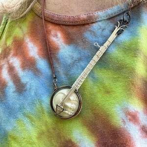 Puede incluir: Un collar con colgante de banjo con una cara color crema y cuerdas y mástil plateados. El colgante está sujeto a un cordón de cuero marrón. El fondo es una camiseta sin mangas teñida anudada en tonos azules, verdes, amarillos y marrones.