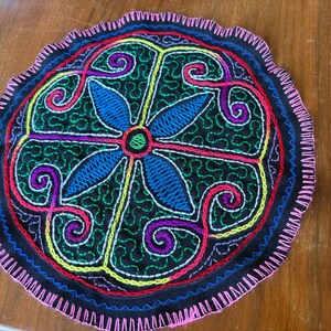 Puede incluir: Un posavasos circular de tela negra con bordados multicolores vibrantes. El diseño incluye una flor central azul de cuatro pétalos, rodeada de intrincados patrones cosidos en verde, amarillo, morado y rojo con un borde de puntada festón rosa.