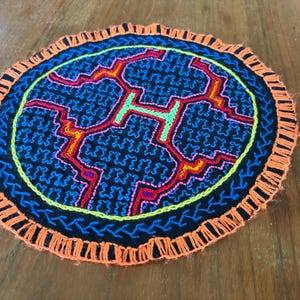 May include: A circular fabric coaster featuring a black base with intricate blue, red, yellow, and green geometric embroidery. The edge is finished with a decorative orange fringe border, creating a textured and colorful circular textile piece.