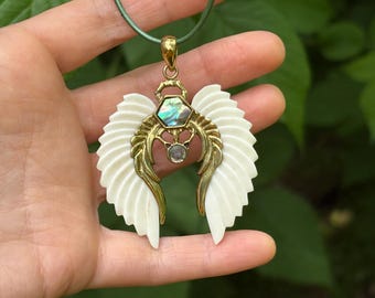May include: A pendant necklace with a wing design. The pendant has cream-coloured wings, gold-coloured metal accents, and an iridescent stone. The necklace hangs from a green cord.