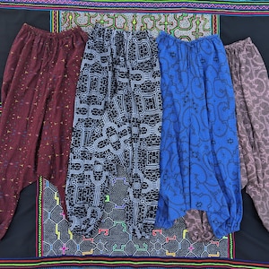 Puede incluir: Cuatro pantalones harén estampados en varios colores: burdeos, blanco y negro, azul y malva. Los pantalones tienen cinturillas y puños elásticos. El fondo es un textil negro con diseños geométricos coloridos.