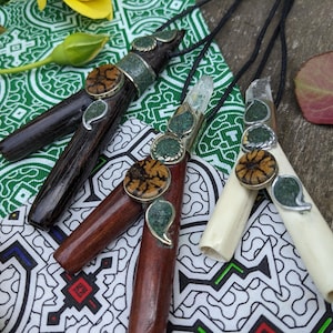 Puede incluir: Primer plano de tres colgantes hechos a mano con elementos de madera, hueso y cristal. Cada colgante presenta detalles únicos de piedra y metal, con un cordón negro. Los colgantes descansan sobre tela estampada.
