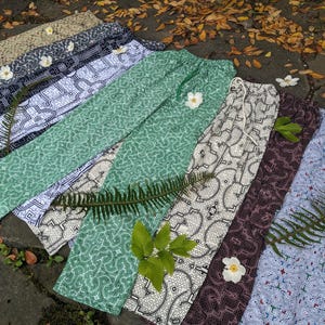 Puede incluir: Una colección de seis pares de pantalones con diferentes patrones abstractos en tonos de verde, blanco, negro y azul. Los pantalones están dispuestos sobre una superficie con hojas y flores.