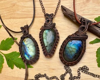 May include: Three handcrafted labradorite pendants with copper wire wrapping. Each pendant features a polished, iridescent gemstone in shades of blue, green, and gold. The pendants are suspended from a black leather cord and copper chain.