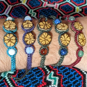 Può includere: Cinque braccialetti macramè fatti a mano in vari colori, ognuno con accenti floreali in legno e perline di pietre preziose. I braccialetti sono turchese, viola, grigio, blu e bordeaux. I braccialetti sono indossati al polso.