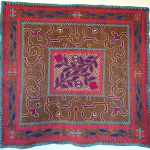 May include: A square brown textile featuring intricate embroidery in shades of pink, blue, and purple. The center displays a floral motif surrounded by geometric patterns and a decorative border with a bright turquoise stitched edge finish.