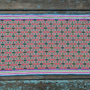 Puede incluir: Textil rectangular cosido a mano con un patrón geométrico repetitivo en rojo, verde y blanco. El borde presenta una franja azul, con un ribete blanco, morado y verde. Colocado sobre una superficie de madera desgastada pintada de azul.