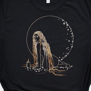 Puede incluir: Camiseta negra con una ilustración de contorno dorado de una mujer sentada con su largo cabello fluyendo a su alrededor. La mujer está rodeada de un círculo de estrellas y una vela está frente a ella.