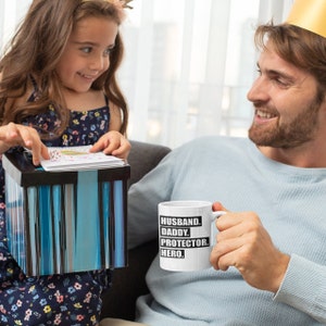 May include: A white ceramic mug with black text that reads "Husband. Daddy. Protector. Hero." A young girl is giving a gift to a man holding the mug.