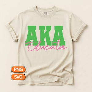 May include: Cream-colored t-shirt with the text "AKA Educator" in green and pink. The word "AKA" is in bold, block letters, and "Educator" is in a cursive font. The shirt has rolled-up sleeves.