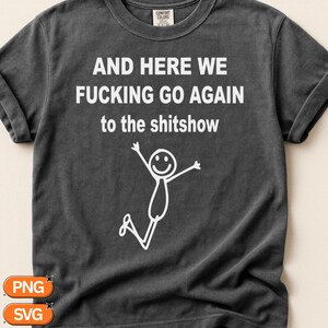 Op de afbeelding: Een antracietgrijs T-shirt met korte mouwen en witte tekst die luidt AND HERE WE FUCKING GO AGAIN to the shitshow, vergezeld van een eenvoudige witte lijntekening van een stokfiguurtje dat springt met opgeheven armen en een lachend gezicht.