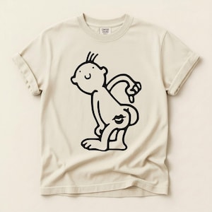 Pode incluir: Camiseta creme com uma figura de desenho animado preta com uma marca de beijo na parte de trás. A figura é desenhada em um estilo simples e divertido. A camisa tem mangas enroladas e gola redonda.