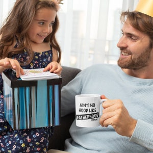 Op de afbeelding: Een witte koffiemok met zwarte tekst die "Ain't no hood like fatherhood #dadlife" zegt. Een man houdt de mok vast en glimlacht naar een jong meisje.