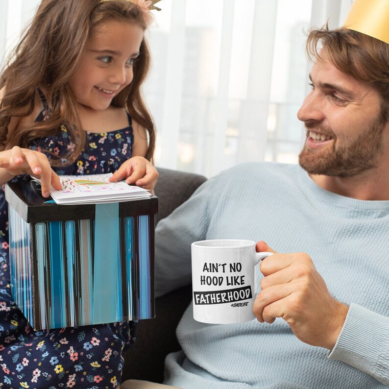 Op de afbeelding: Een witte koffiemok met zwarte tekst die "Ain't no hood like fatherhood #dadlife" zegt. Een man houdt de mok vast en glimlacht naar een jong meisje.