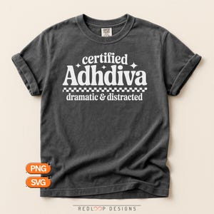 May include: A charcoal grey short-sleeve t-shirt featuring white text that reads certified Adhdiva with small star icons, a checkered line, and the words dramatic & distracted below. The shirt has a crew neckline and a relaxed fit.