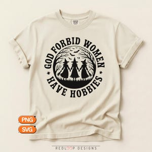 Może przedstawiać: Kremowy t-shirt z krótkim rękawem z czarną, okrągłą grafiką przedstawiającą trzy czarownice trzymające się za ręce pod koroną drzew z nietoperzami. Napis GOD FORBID WOMEN HAVE HOBBIES jest umieszczony pogrubioną czcionką wokół ilustracji.