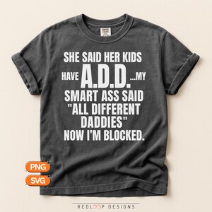 May include: A charcoal gray short-sleeve t-shirt featuring white text that reads: SHE SAID HER KIDS HAVE A.D.D....MY SMART ASS SAID "ALL DIFFERENT DADDIES" NOW I'M BLOCKED. The shirt has a crew neckline and a small tag at the inner collar area.
