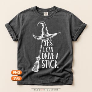 May include: A charcoal gray short-sleeve t-shirt featuring a white graphic of a witch hat atop a broomstick. The text reads, YES I CAN DRIVE A STICK. The shirt has a crew neckline and a vintage-style wash, with a Comfort Colors brand label visible.