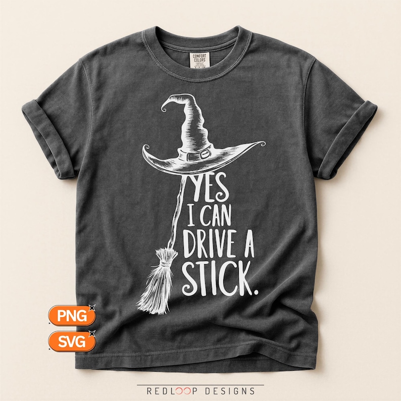 May include: A charcoal gray short-sleeve t-shirt featuring a white graphic of a witch hat atop a broomstick. The text reads, YES I CAN DRIVE A STICK. The shirt has a crew neckline and a vintage-style wash, with a Comfort Colors brand label visible.