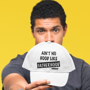 Op de afbeelding: Witte baseballpet met zwarte tekst die "Ain't no hood like fatherhood #dadlife" zegt.