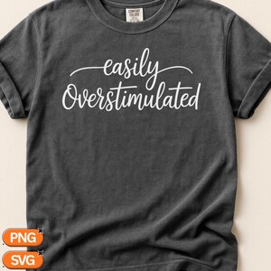 Puede incluir: Una camiseta de manga corta color gris carbón con el texto blanco "easily Overstimulated" impreso en una combinación de fuente cursiva y sans-serif en el pecho. La prenda tiene cuello redondo y una etiqueta de la marca Comfort Colors.