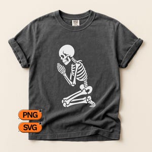 May include: Dark grey t-shirt with a white skeleton kneeling in prayer. The skeleton's hands are clasped, and the design is centered on the shirt. The shirt has rolled-up sleeves and a Comfort Colors tag.