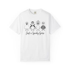 May include: White t-shirt featuring four cartoon geese dressed in Halloween costumes: a ghost, a vampire, a jack-o'-lantern, and a skeleton. The phrase "Just a Spooky Goose" is printed below the geese in a script font. The shirt has a crew neck.