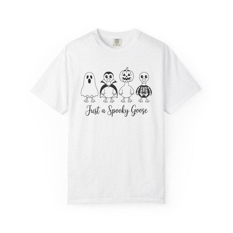 May include: White t-shirt featuring four cartoon geese dressed in Halloween costumes: a ghost, a vampire, a jack-o'-lantern, and a skeleton. The phrase "Just a Spooky Goose" is printed below the geese in a script font. The shirt has a crew neck.