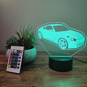 May include: A 3D LED night light shaped like a car with a remote control. The light is on and is a bright green color. The remote control has 16 different color options.