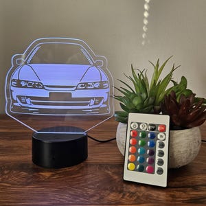 May include: A car-shaped LED night light with a black base and a remote control. The light emits a blue glow, and the remote has multiple color options. A small potted plant sits nearby on a wooden surface.
